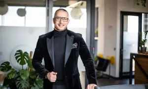 Mężczyzna w czarnym garniturze i okularach stoi w nowoczesnym biurze, uśmiecha się.