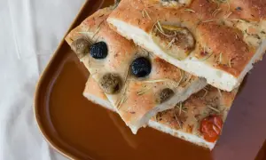 Focaccia Pomodoro mit Oliven und Rosmarin auf brauner Platte.