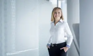 Uśmiechnięta kobieta w białej koszuli i czarnych spodniach, stojąca w nowoczesnym biurze.