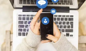 Dłonie trzymające smartfon nad klawiaturą laptopa. Ikony komunikatorów unoszą się nad ekranem. Chcesz wiedzieć, jak sprawdzić kiedy ktoś był aktywny na Messengerze?