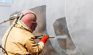 Pracownik w kombinezonie i masce piaskuje duży zbiornik. Sprawdza jakie pozwolenia na piaskowanie są potrzebne do tej pracy.