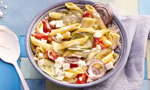 Schüssel mit griechischem Nudelsalat: Penne, Gurken, Tomaten, rote Zwiebeln und Feta in cremiger Soße.
