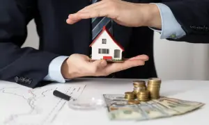 Mani che proteggono una casa modello, con grafici, soldi e monete. L'idea di comprare casa per affittarla è ben rappresentata.