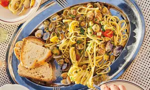 En stor silverbricka med pasta vongole, musslor, körsbärstomater och vitlöksbröd. En hand tar en gaffel med pasta.