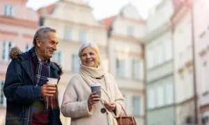 Uśmiechnięta para seniorów z kawą w rękach spaceruje po urokliwej uliczce. Marzą o podróżach, może o wycieczkach dla seniorów samolotem?