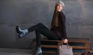 Modna dziewczyna w czarnych rurkach i swetrze pokazuje, jak nosić sztyblety damskie. Stylizacja z szarą czapką i torebką.