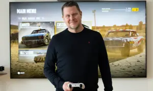 Carolin Wingefors ler mot kameran med en spelkontroll i handen, framför en TV som visar spelets huvudmeny med racerbilar.
