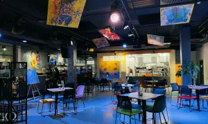 Wnętrze Oria Magic House z kolorowymi obrazami na suficie i ścianach. Stoliki i krzesła tworzą przytulną atmosferę, idealną do przeglądania menu.