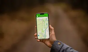 Dłoń trzyma smartfon z mapą i trasą. Aplikacja do planowania podróży pokazuje ścieżkę przez tereny zielone i wodne.