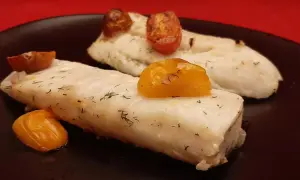 Deux pavés de dos de merlu tendres, parsemés d'aneth et surmontés de tomates cerises rôties, sur une assiette noire.