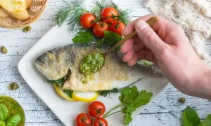 Eine goldbraune Dorade in der Pfanne, garniert mit frischen Kräutern, Kirschtomaten und Zitronenscheiben. Eine Löffelspitze grüner Sauce wird auf den Fisch gegeben.