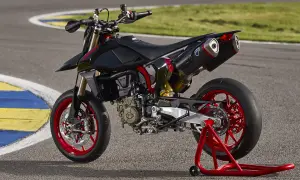 Czarny motocykl **hypermotard** z czerwonymi felgami i akcentami, stojący na czerwonym stojaku na torze wyścigowym.