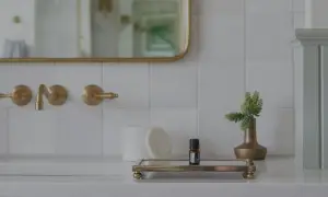 Elegancka, biała łazienka ze złotymi dodatkami. Na blacie stoi mała buteleczka olejku i złoty wazonik z zieloną rośliną.
