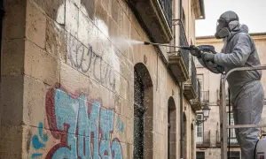 Trabajador con traje protector realiza limpieza de fachada con hidrolimpiadora, eliminando grafitis de un muro de piedra.