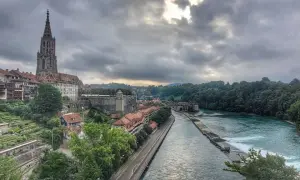 Blick von der Besucherplattform über Bern: Münster, Aare und grüne Hänge unter dramatischem Himmel.