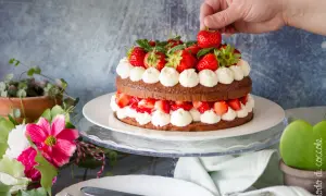 Una deliziosa torta con fragole e cioccolato, decorata con panna montata e fragole fresche. Una mano aggiunge l'ultima fragola in cima.