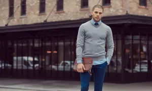 Ein Mann mit Stil, der sein **Outfit zusammenstellen Herren** perfektioniert: grauer Pullover, hellblaues Hemd, blaue Hose und eine braune Ledertasche.
