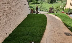 Sentiero in cemento stampato spessore minimo, con finitura effetto pietra, fiancheggiato da prato verde e muro in pietra grezza.