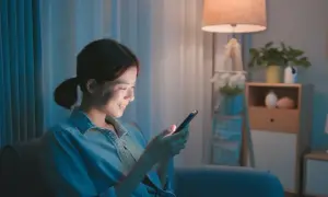Uśmiechnięta kobieta z telefonem w ręku, siedząca w przytulnym, oświetlonym lampą pokoju.