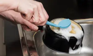 Ręka szoruje czarną patelnię z pianą, używając niebieskiej szczotki. Boraks co to? To naturalny środek czyszczący.