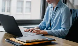 Osoba w niebieskiej koszuli pracuje przy laptopie, tworząc skrypty VBA. Na biurku leżą notatniki.
