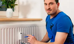 Fachowiec w niebieskim kombinezonie sprawdza odczyty na podzielnikach ciepła zamontowanych na grzejniku.