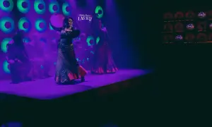 Dos bailarinas de flamenco, una con un tambor, aprenden a bailar sevillanas en un escenario con luces moradas y verdes.