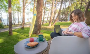 Kobieta pracuje na laptopie przy stoliku z kawą i croissantem. W tle widać morze i domki z wyjściem na plażę.