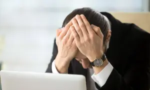 Pracownik w garniturze z rękami na twarzy, przy laptopie. Wygląda na przygnębionego, być może z powodu odpowiedzialności finansowej pracownika za błędy.