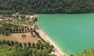 Vue aérienne d'un magnifique lac aux eaux turquoise, idéal pour la baignade. Des plages aménagées bordent la rive, entourées de verdure et d'arbres.