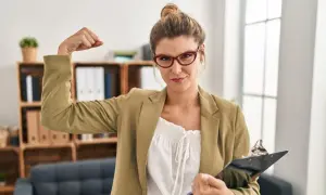 Kobieta w okularach pokazuje biceps, symbolizując, jak być silnym psychicznie. W ręku trzyma teczkę.