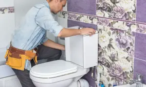 Instalator w pasie z narzędziami montuje biały stelaż podtynkowy do toalety. Łazienka ozdobiona jest fioletowymi płytkami z kwiatowym wzorem.