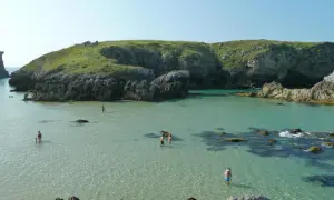 Gente disfrutando del agua cristalina en la costa de Llanes, con acantilados verdes y rocosos de fondo.