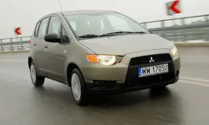 Złoty Mitsubishi Colt 2006 na mokrej drodze, światła włączone, w tle znaki drogowe.