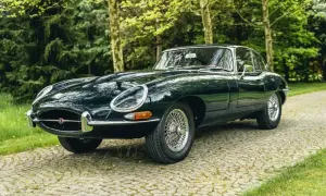Klasyczny, ciemnozielony Jaguar E-Type na brukowanej drodze, otoczony drzewami.