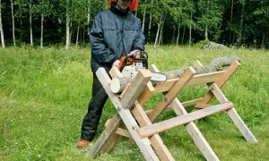 Man med motorsåg använder en hemmagjord sågbock för att kapa ved i en grönskande skogsmiljö.
