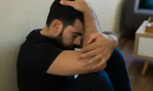 Mężczyzna siedzi przy ścianie, obejmując kolana. Wygląda na przygnębionego, jakby przeżywał jakiś trudny moment, może nawet kac moralny.