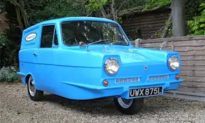 En blå Reliant bil, en trehjulig klassiker, står parkerad på grus.