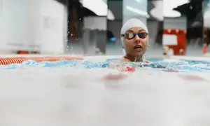 Kobieta w czepku i okularach pływackich wynurza głowę z wody w basenie.