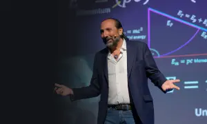 Nassim Haramein, con gestos amplios, explica conceptos de física ante una pantalla con fórmulas como E=mc².