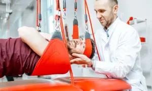 Fizjoterapeuta w białym fartuchu pomaga pacjentowi w rehabilitacji kręgosłupa szyjnego na czerwonej macie.