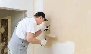 Mężczyzna w ogrodniczkach i czapce maluje ścianę wałkiem. Lampa do malowania ścian jest niezbędna do precyzyjnego wykończenia.