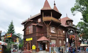 Drewniana architektura na Krupówkach, z charakterystycznymi zdobieniami i wieżyczkami. W tle widać ludzi spacerujących po tętniącej życiem ulicy.