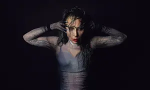 Lady Gaga w siateczkowej bluzce z długimi rękawami i tatuażami, zakrywająca twarz dłońmi.