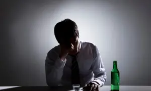 Mężczyzna w białej koszuli i krawacie, z ręką na czole, siedzi przy stole. Obok niego stoi zielona butelka i szklanka. Wygląda na to, że zmaga się z zatruciem alkoholowym.