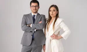 Para w garniturach, mężczyzna z rękami skrzyżowanymi na piersi, kobieta z ręką na biodrze.