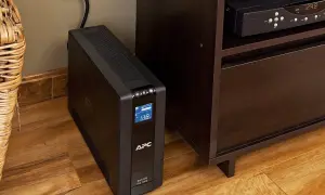 Czarny UPS APC stoi na drewnianej podłodze obok mebla. Jaki UPS, chroni sprzęt przed zanikiem prądu.