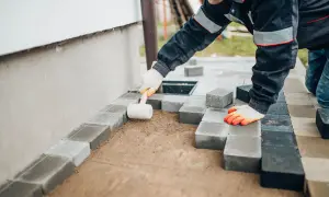 Pracownik w rękawicach układa kostkę brukową przy elewacji, używając gumowego młotka do precyzyjnego dopasowania.