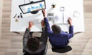 Två personer vid ett skrivbord analyserar data på en skärm, ett exempel på it-system. En person i rullstol skriver på tangentbordet, den andra pekar på diagrammet.