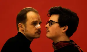 Karaś/Rogucki - Marianna. Dwóch mężczyzn zapatrzonych w siebie na czerwonym tle. Jeden ma plaster na nosie.